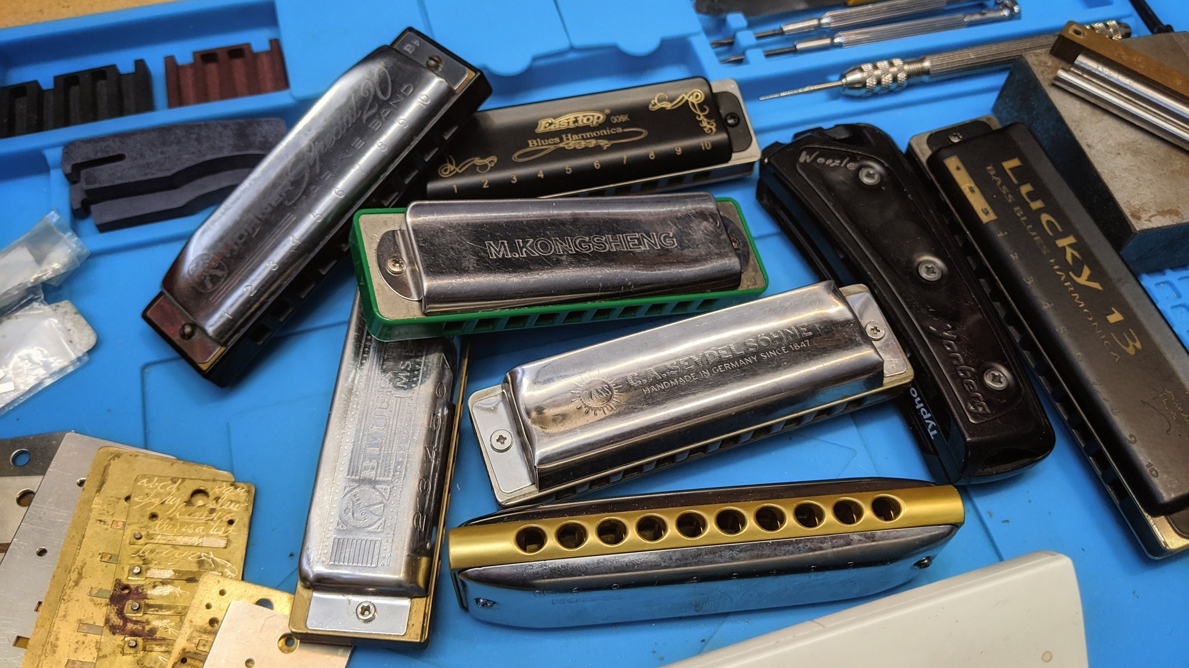 A variety of harmonicas from different brands, along with reed-plates, and miscellaneous tools on a blue rubber work pad.